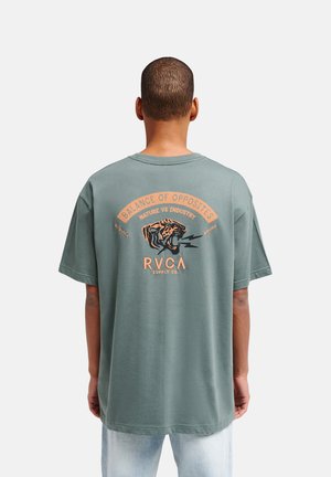Homme de dos portant un t-shirt vert avec un tigre rugissant et le texte « Balance of Opposites Nature vs Industry RVCA » dans le dos.