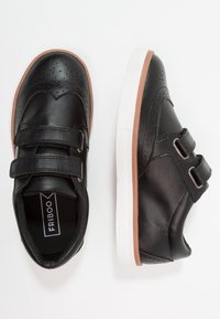 Chaussures en cuir noir avec des détails perforés, des bandes Velcro, une semelle blanche et des accents marron. Bout arrondi et texture lisse.