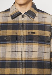 Károvaná flanelová košile v béžové, námořnické a modré barvě. Má zipové zapínání, dvě přední kapsy a značkovou nášivku na levé straně. Texturovaná látka.