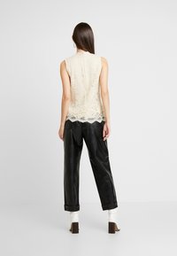 Kvinna med långt mörkt hår, klädd i en ärmlös beige spetsblus, svarta strukturerade byxor och vita klackade ankelskor, står mot en vit bakgrund.