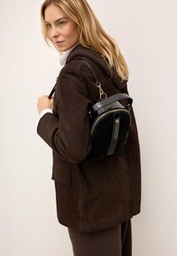 Čierny lakovaný a semišový batoh s zaobleným tvarom, zipsovým uzáverom a zlatými kovovými doplnkami, nosený na hnedej bunde.