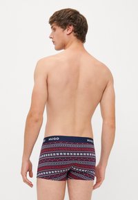 Junger Mann mit kurzen braunen Haaren, oben ohne, trägt gemusterte dunkelblaue, rote und weiße Boxershorts mit "HUGO"-Logo am Bund, mit dem Rücken zur Kamera.
