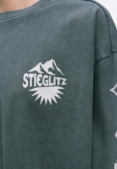 Zelená mikina s jemnou textúrou, s bielym grafickým dizajnom pohoria a textom "STIEGLITZ" na prednej strane. Vysoký okrúhly výstrih, uvoľnený strih.