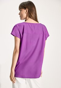 Blusa viola a maniche corte, con scollatura rotonda e taglio dritto. Il tessuto sembra leggero e ha una texture liscia.