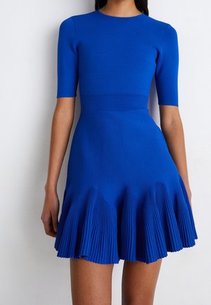 Femme portant une robe bleue cintrée à manches courtes, taille côtelée et jupe évasée plissée se terminant au-dessus des genoux.