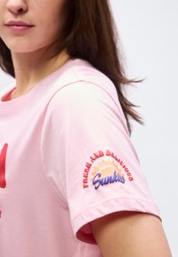 Γυναίκα που φοράει ανοιχτό ροζ μπλουζάκι με το κείμενο "Φρέσκο και νόστιμο Sunkiss" και γραφικό του ήλιου στο μανίκι, κομμένη στο πρόσωπο και το χέρι.