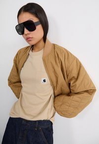 Veste bomber matelassée beige avec un t-shirt beige arborant un logo sur la poche poitrine. Grandes lunettes de soleil noires. Jeans bleu marine avec des poches traditionnelles.