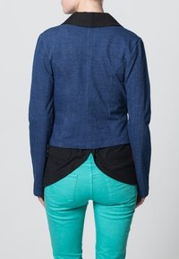 Blå denimjacka med svart krage, med en figurnära design och kort längd, bärs över en svart topp och matchas med gröna byxor.