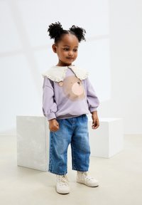 Felpa lilla con grafica orso e scollo con colletto in pizzo bianco, abbinata a jeans a gamba larga blu e sneakers bianche alte.