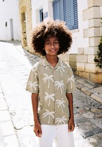 Camicia a maniche corte color verde chiaro con motivi di palme bianche, abbinata a pantaloni bianchi, indossata da un bambino con capelli ricci e voluminosi.