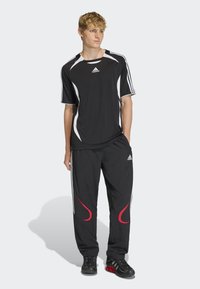 Jauns vīrietis stāv, ģērbies melnā Adidas sporta apģērbā ar baltajiem akcentiem un sarkanajiem detaļām biksēs, melnās un sarkanās sporta kurpēs.