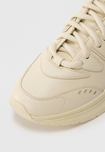 Αθλητικό παπούτσι από εκρού δέρμα με υφασμένους τόνοι, διαθέτει κορδόνια στο ίδιο χρώμα και ελαφρώς υφασμένο λαστιχένιο πέλμα. Καμπύλες γραμμές σχεδίασης σε όλο το παπούτσι.