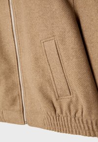 Section d'une veste en laine beige montrant une poche latérale avec piqûres en bordure, une fermeture éclair argentée et un détail d'ourlet élastiqué.