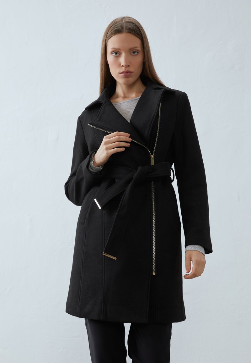 Svart ullblandingsfrakk med belte i midjen, asymmetrisk glidelås og notched krage. Glatt tekstur og minimalistisk design.