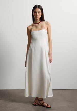 Donna con un vestito midi bianco senza maniche, collana di perline rosse e sandali bassi marroni con cinturini, in piedi contro uno sfondo semplice.