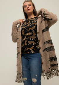Beige Strickjacke mit schwarzen Streifen und Fransen. Darunter trägt sie ein schwarzes Shirt mit goldenen Leopardenmustern. Kombiniert mit blauen, zerrissenen Jeans.