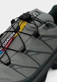 Lys grå joggesko med teksturert overdel, svarte elastiske snorer, en gul aksent og geometriske designelementer. Har vanntett Gore-Tex.