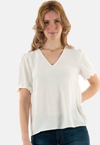 Blouse blanche à manches courtes avec un col en V, présentant des détails festonnés sur les manches, fabriquée à partir d'un tissu texturé. Associée à un jean bleu marine.