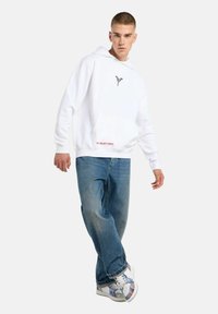 Witte hoodie met een voorzak, voorzien van een klein zwart logo, gecombineerd met lichtblauwe baggy jeans en kleurrijke, gedessineerde sneakers.