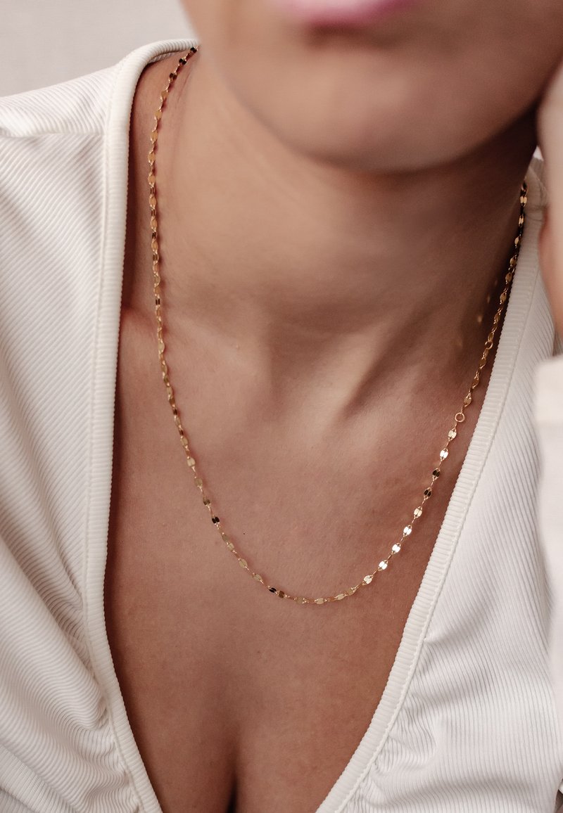 Złoty łańcuszek z teksturowanymi, wydłużonymi ogniwami, spoczywający na nagiej skórze, na tle jasnej, żebrowanej bluzki z dekoltem w kształcie litery V.