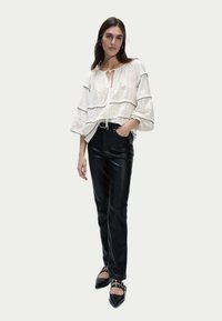 Blusa bianca con texture e rifiniture nere, dal taglio morbido e dettaglio a fiocco sul collo, abbinata a pantaloni in pelle neri a vita alta e scarpe a punta.