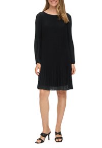 Schwarzes, plissiertes Kleid mit langen Ärmeln, Rundhalsausschnitt und knielanger Saumlänge. Getragen mit schwarzen High-Heels. Glatte Textur.