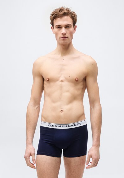 Νεαρός άνδρας με σγουρά μαλλιά που φοράει μποξέρ Polo Ralph Lauren σε σκούρο μπλε, στέκεται μπροστά σε ένα απλό ανοιχτό γκρι φόντο.