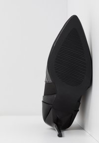 Chaussure à talon noir avec un bout pointu, en cuir texturé, semelle rainurée et design élégant. Le talon est fin et angulaire.