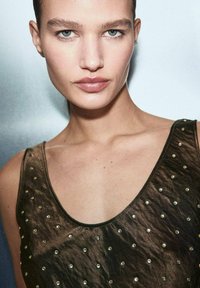 Smeđa bezrukavna majica s dubokim izrezom, izrađena od teksturirane tkanine i ukrašena dekorativnim metalnim zakovicama ravnomjerno raspoređenim po prednjem dijelu.