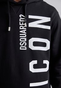 Sudadera con capucha negra con grandes textos blancos "DSQUARED2" e "ICON". El material es algodón suave, con cordones con puntas metálicas y un acabado texturizado.