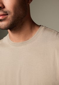 Nahaufnahme des unteren Gesichts und Halses eines Mannes, der ein beiges T-Shirt mit Rundhalsausschnitt vor einem neutralen Hintergrund trägt.