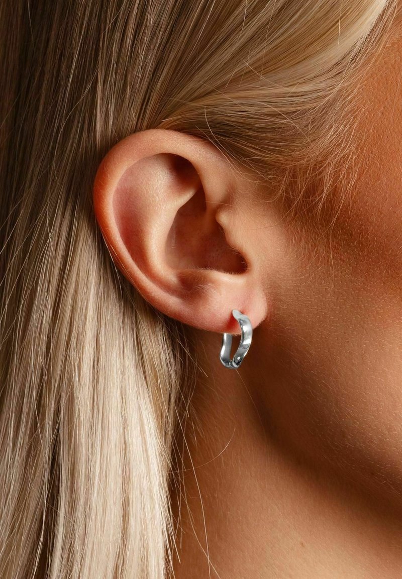 Silver hoop earring displayed on a model’s ear, featuring a smooth, polished surface and a circular, slightly elongated design.