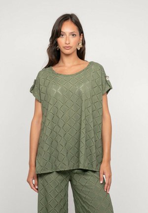 Femme portant un haut en maille vert olive et un pantalon assorti avec un motif en losange, debout devant un fond uni clair.