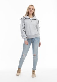 Lichtgrijze sweatshirt met een rits-halslijn, oversized kraag en geribde zoom, gecombineerd met gewassen lichtblauwe jeans en beige hoge sneakers.