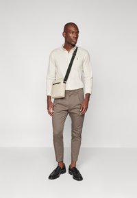Camisa beige de botones, pantalones marrones ajustados, mocasines negros y un pequeño bolso bandolera con cremallera. El fondo es blanco liso.