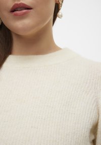Maglione crema lavorato a maglia con scollatura arrotondata e texture a coste; indossato con orecchini dorati caratterizzati da un grande accento di perla.