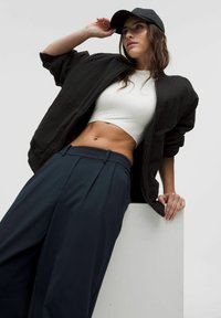 Black bomber jacket, white crop top, and navy wide-leg trousers. The model poses with a casual posture, wearing a black cap.