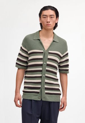 Mand iført en grøn, creme og brun stribet kortærmet strikket skjorte med knapper og mørke bukser, stående mod en ensfarvet baggrund.