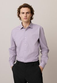 Helllila Langarmshirt mit Button-Down-Kragen, aus glattem Stoff. Kombiniert mit dunklen Hosen, die Seitentaschen haben.