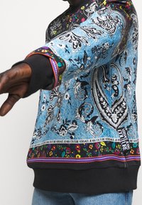 Blå sweatshirt med ett paisleymönster, svarta ribbade manschetter, färgglada blomaccent på nederkanten och logodetaljer längs den nedre bandet.