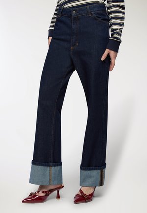 Jeans a gamba larga in denim blu scuro con vita alta, caratterizzati da un risvolto in denim chiaro a contrasto. Abbinati a scarpe con tacco rosso lucido.
