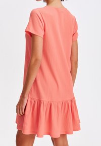 Vestido corto color coral de manga corta con tejido texturizado, escote redondo y dobladillo con volantes. El diseño presenta un corte holgado y un estilo casual.