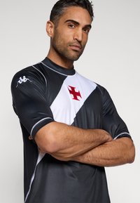 Kratka športna majica z rokavi v črno-beli barvi, s centralnim rdečim križem, izdelana iz gladkega, raztegljivega materiala.