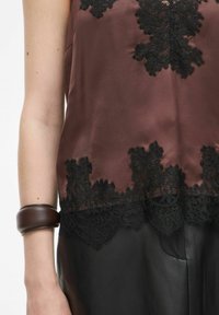 Brązowa satynowa bluzka z czarną koronką na dolnej krawędzi. Koronka ma skomplikowane wzory. Częściowo widoczne są ciemne skórzane spodnie.