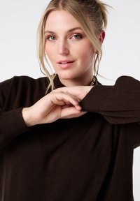 Bruine gebreide trui met een ontspannen pasvorm en geribbelde manchetten, met een ronde halslijn en een gestructureerde stof. Handen rusten op de mouwen.