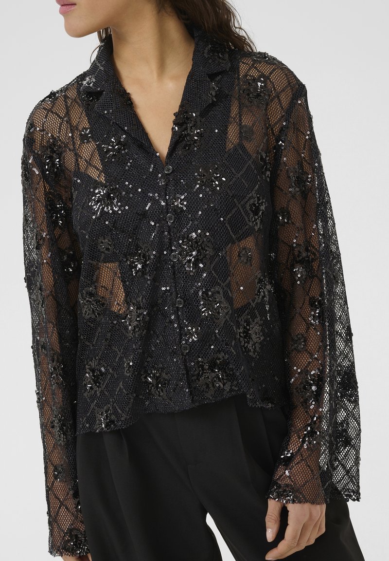 Bluză neagră transparentă, cu un model floral din paiete, cu închidere cu nasturi în față și mâneci lungi cu textură asemănătoare dantelei. Design elegant și ușor.