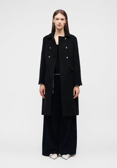 Frau steht und trägt einen langen schwarzen doppelt geknöpften Mantel, weite schwarze Hose, Kettengürtel und weiße spitze Schuhe vor einem einfarbigen Hintergrund.