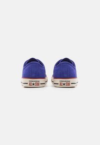 Baskets basses en toile violette avec des semelles en caoutchouc blanc contrastant, arborant la marque "Converse All Star" sur le talon et une couture texturée.