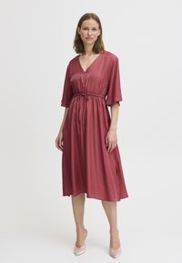 Robe rose avec un décolleté en V, des manches courtes, une taille élastique et une jupe évasée fluide. Tissu lisse avec un léger éclat.