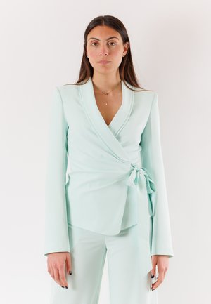 Donna con capelli lunghi e castani che indossa un blazer avvolgente azzurro pallido annodato in vita e pantaloni abbinati su uno sfondo semplice.